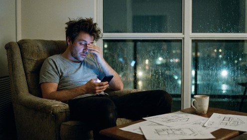 A tired man with messy hair sitting in a dimly lit living room at night. He is rubbing his eyes while looking at a glowing smartphone screen. In the foreground, architectural blueprints are scattered on a table next to a coffee mug. Outside the large window, it is raining, with city lights blurred in the background. Cinematic, moody atmosphere.